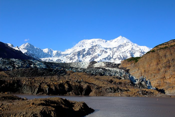 Mount St. Elias rises above the head of Taan Fjord, with Tyndall Glacier in the background. The glacier is covered with debris from a recent landslide. From sea level at this point to the summit of St. Elias is just 11 miles horizontally and 18,000 feet vertically.